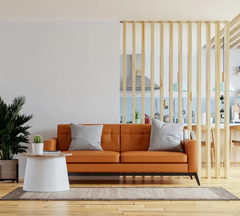 Pomarańczowa, tapicerowana sofa z dwiema szarymi poduszkami ustawiona przed pionowymi, jasnymi lamelami dzielącymi przestrzeń, obok biały okrągły stolik z małym kaktusem i duża roślina doniczkowa, w tle widoczna jasna, otwarta kuchnia.