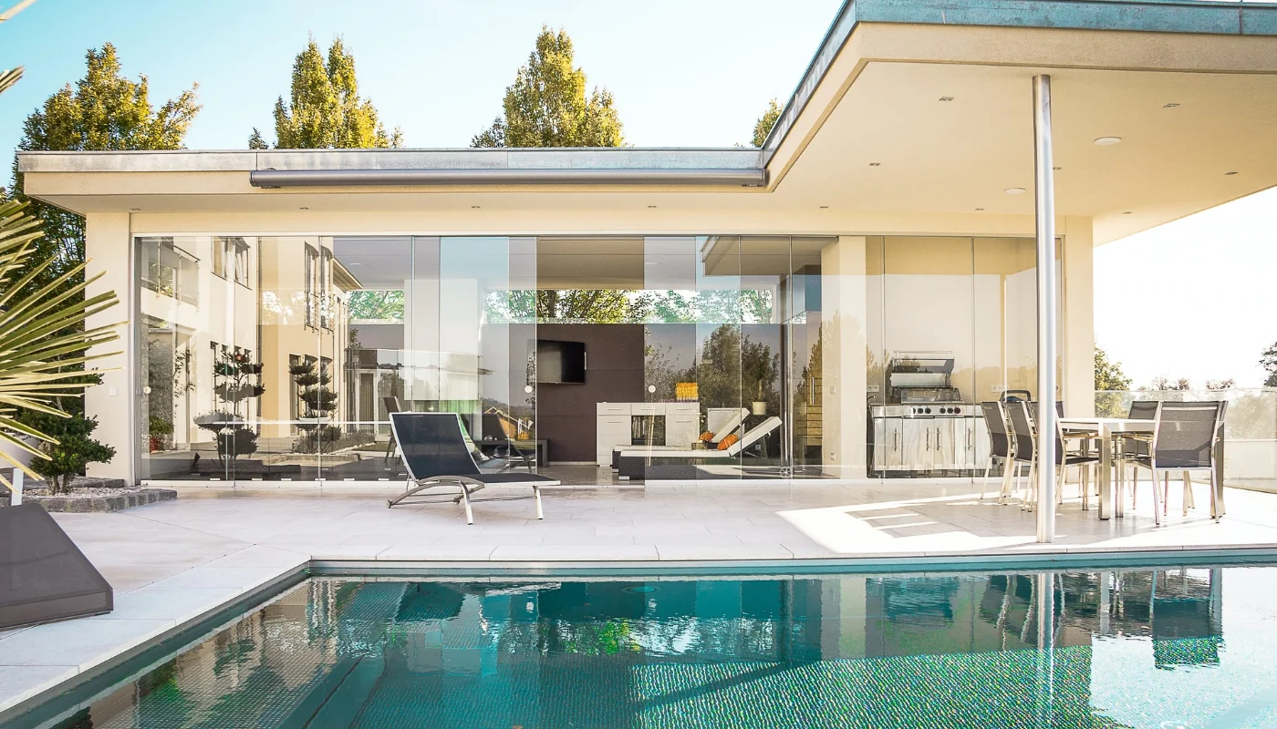 Nowoczesny, parterowy dom z przeszkloną elewacją otwarty na jasny taras z leżakiem i stołem jadalnym przy wbudowanym grillu, a na pierwszym planie prostokątny basen odbijający budynek i drzewa.