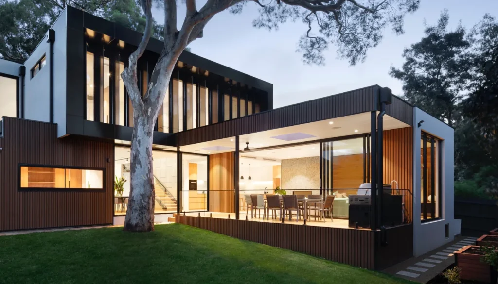 Nowoczesny dwupiętrowy dom wieczorem z dużymi przeszkleniami i ciemnymi ramami, elewacją z poziomych drewnianych paneli, przeszklonym zadaszonym tarasem z jadalnią i grillem, widocznym ciepłym wnętrzem oraz dużym drzewem i zielonym trawnikiem na pierwszym planie.
