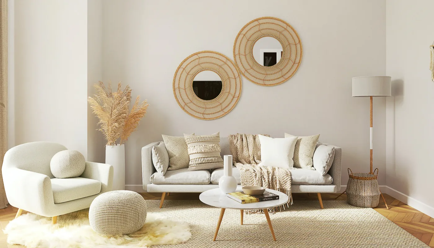 Przytulny, neutralny salon w odcieniach beżu z jasnoszarą sofą ozdobioną teksturowanymi poduszkami i kocem, fotelem i pufem po lewej, niskim okrągłym stolikiem kawowym na środku, dwiema plecionymi okrągłymi lustrami na ścianie, wysokim wazonem z trawami pampasowymi po lewej oraz stojącą lampą i plecionym koszem po prawej.