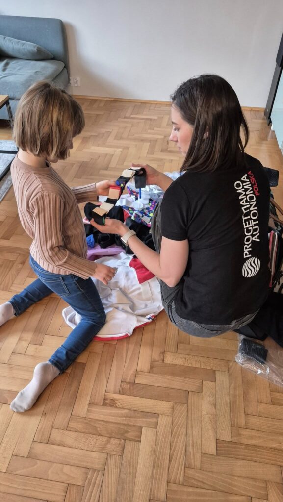 Kobieta i dziecko klęczą na jasnym jodełkowym parkiecie w salonie; kobieta pokazuje dziecku kolorowy zestaw pasków i materiałów rozłożonych na kocu obok kanapy.