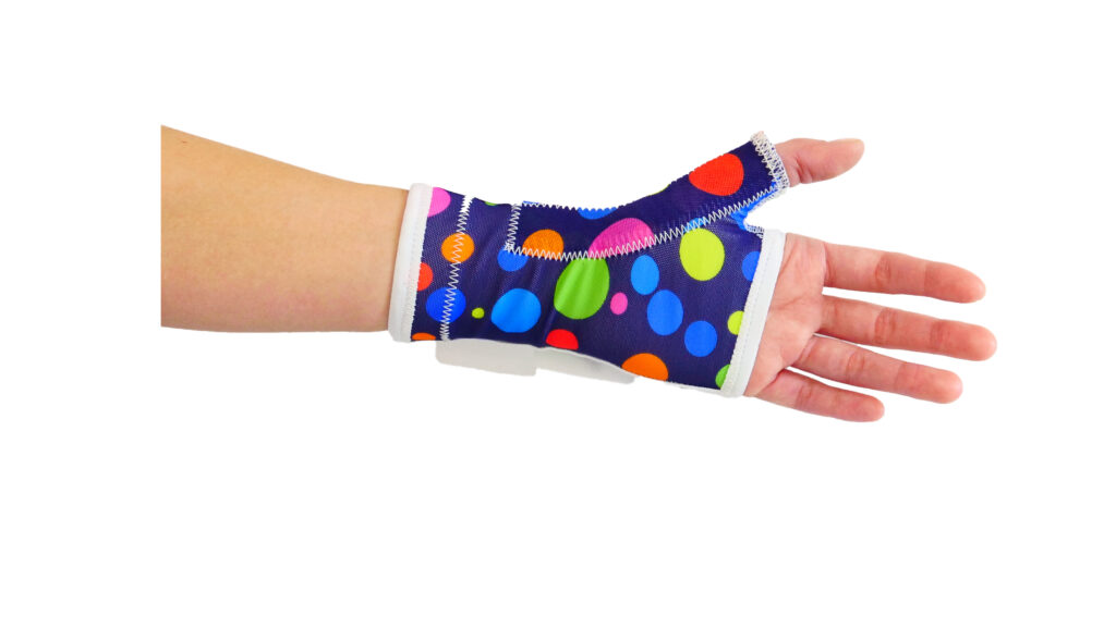 Otwarta dłoń i przedramię w kolorowej ortezie nadgarstka z wycięciem na kciuk — granatowy stabilizator w duże wielobarwne kropki z białym obszyciem na białym tle.