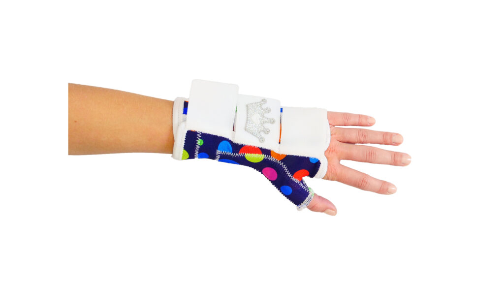 Dłoń i przedramię w ortezie nadgarstka z granatowym materiałem w duże kolorowe kropki, białymi rzepami i podpórką na kciuk, ozdobione srebrną naszywką w kształcie korony, na białym tle.