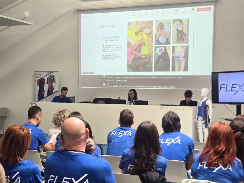 Publiczność w niebieskich koszulkach z napisem FLEXA siedzi na sali konferencyjnej i słucha trzech prelegentów za stołem przed dużym ekranem projekcyjnym wyświetlającym slajd z fotografiami i tekstem, obok stoi manekin w ortopedycznym ubraniu.