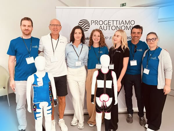 Siedmiu dorosłych w identyfikatorach i firmowych koszulkach stoi uśmiechniętych przed banerem "PROGETTIAMO AUTONOMIA", obok dwóch dziecięcych manekinów ubranych w kolorowe ortopedyczne uprzęże i kombinezony demonstracyjne.