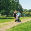 Mężczyzna na elektrycznym wózku inwalidzkim w jasnoniebieskiej koszulce rozmawia i uśmiecha się do idącej obok blondynki w czarnej sukience podczas spaceru po ścieżce w słonecznym parku z drzewami i placem zabaw w tle.