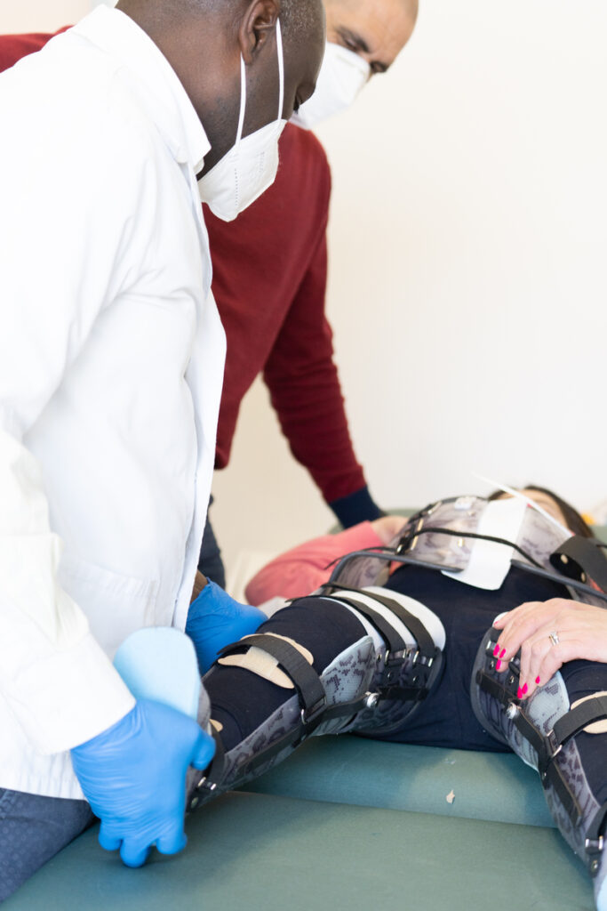 Lekarz w białym fartuchu i niebieskich rękawiczkach dopasowuje ortezę na nodze pacjentki leżącej na stole zabiegowym, obok pochyla się inna osoba w masce.