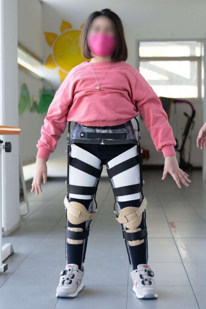 Dziecko w różowej bluzie i różowej maseczce stoi na korytarzu ośrodka rehabilitacyjnego, ma na nogach białe ortezy z czarnymi pasami i podpórkami oraz sportowe buty.