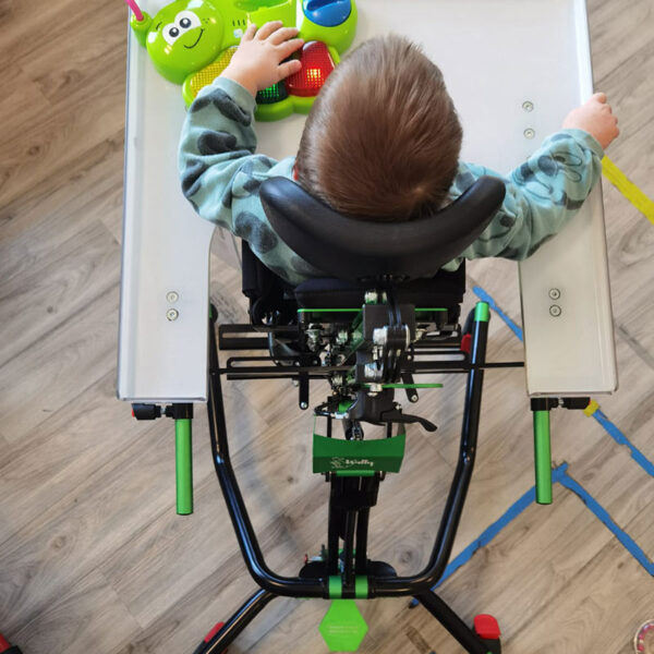 Widok z góry na małe dziecko opierające się w pionizatorze z czarnym zagłówkiem i zielono-czarną ramą, przy białym stoliku bawiące się zieloną interaktywną zabawką na drewnianej podłodze