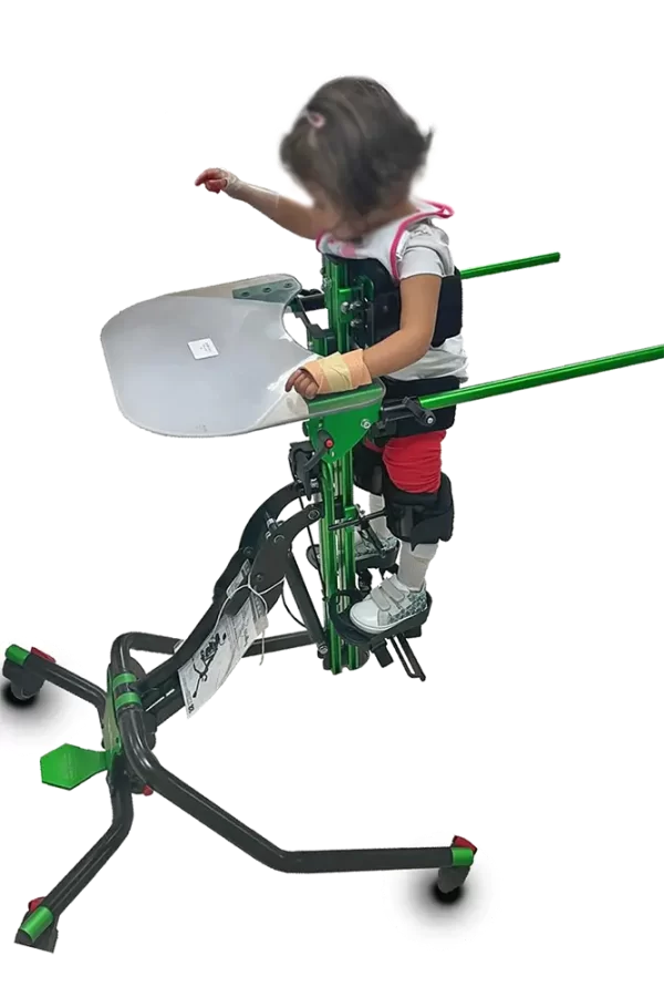 Małe dziecko stoi w zielonej ramie rehabilitacyjnej z przymocowaną półkolistą tacą, przypięte pasami do podtrzymania tułowia i ud, stopy na podpórkach, a konstrukcja ma czarną podstawę na kółkach.