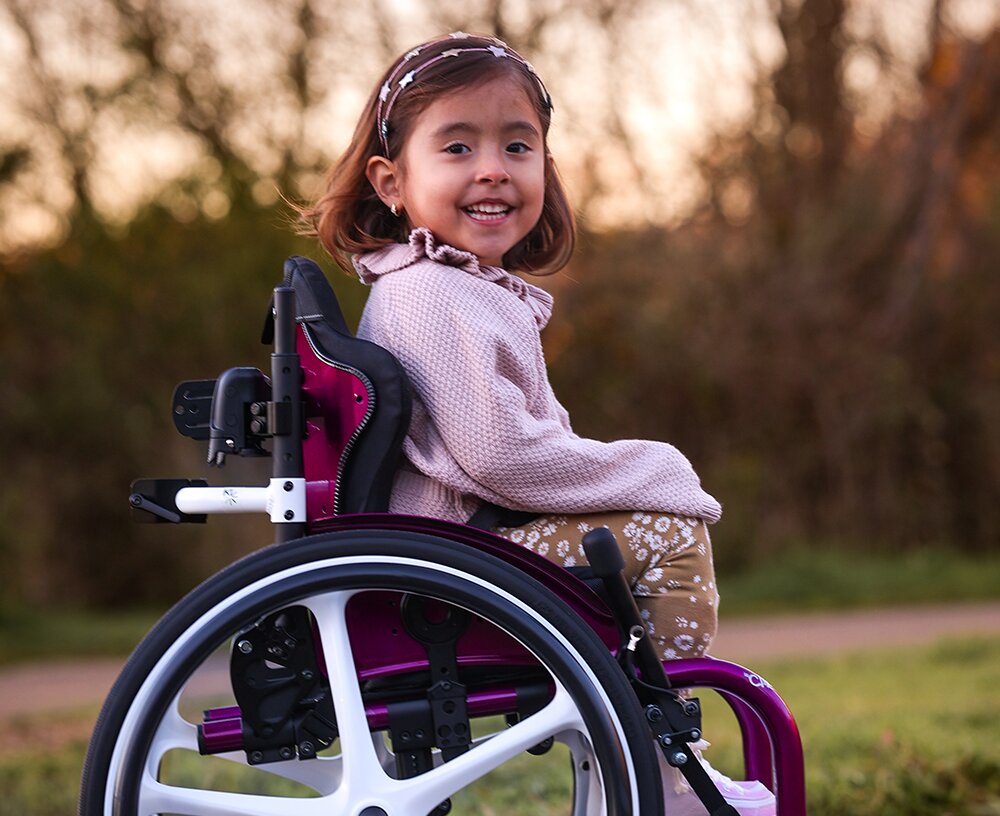 Uśmiechnięta mała dziewczynka w różowym sweterku i opasce na głowie siedzi w fioletowym wózku inwalidzkim na zewnątrz.