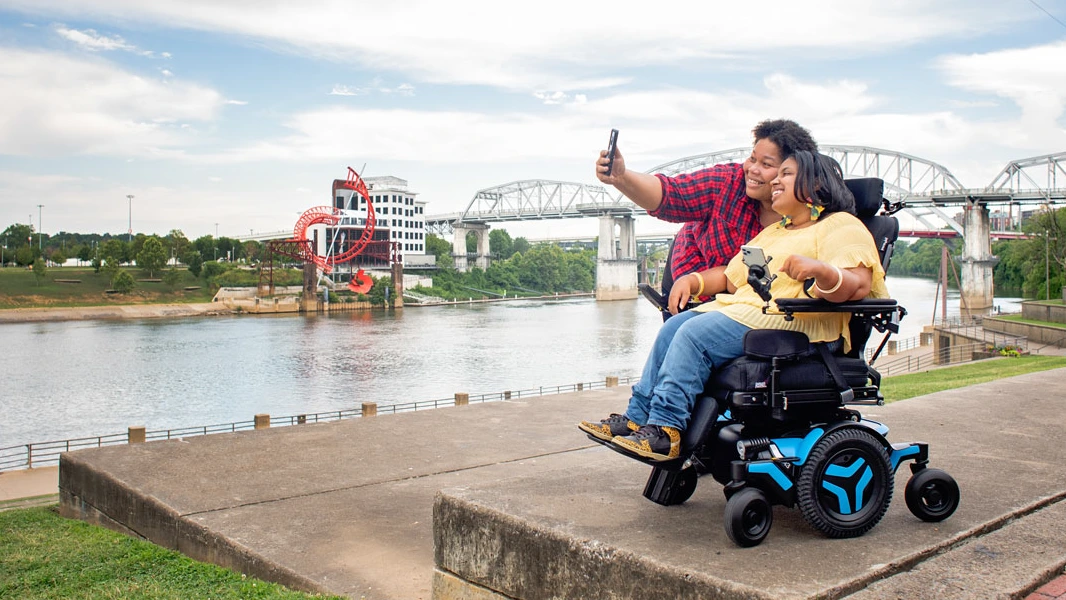 Para uśmiechniętych osób, jedna na elektrycznym wózku inwalidzkim, robią razem selfie na nadbrzeżu z rzeką i mostami w tle.