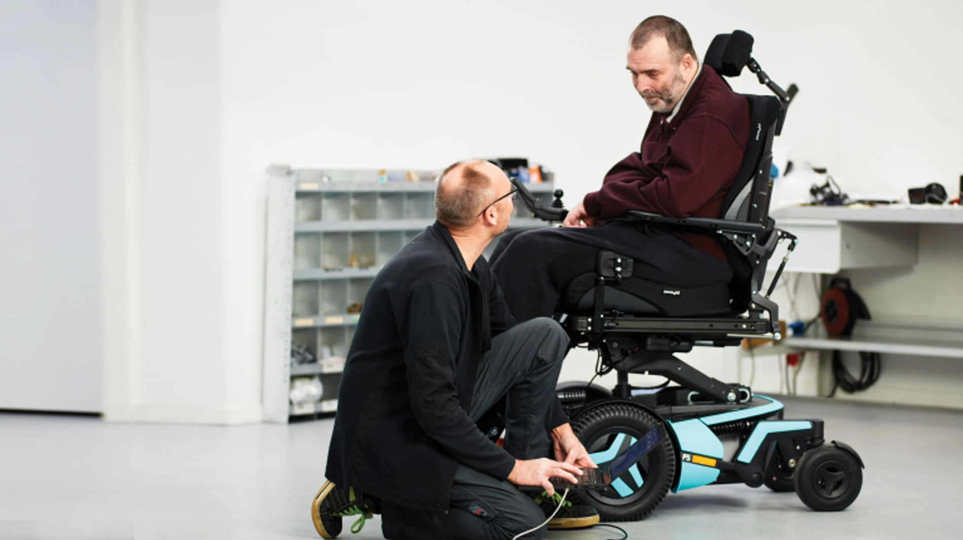 Technik klęczy przy elektrycznym wózku inwalidzkim, podłączając urządzenie diagnostyczne przy kole, a użytkownik siedzi i obserwuje.
