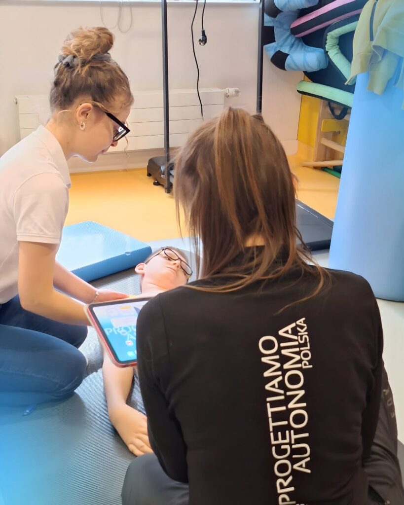 Dwie osoby nad osobą leżącą na macie, jedna trzyma tablet; na czarnej koszulce napis PROGETTIAMO AUTONOMIA.