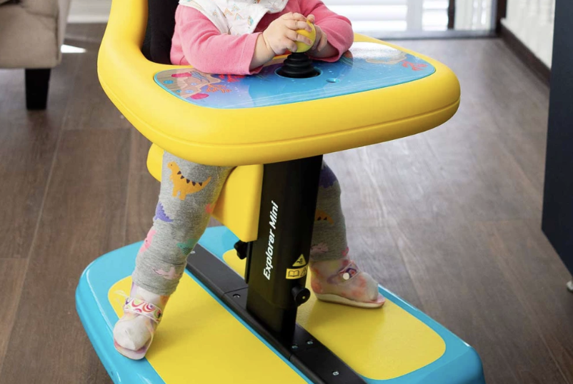 Dziecko w żółto‑niebieskim chodziku trzyma joystick, ma legginsy z dinozaurami i skarpetki, stoi na drewnianej podłodze.