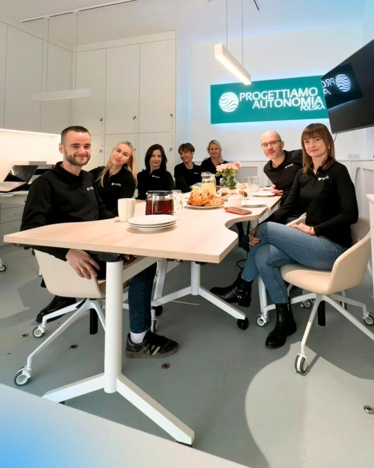 Grupa ośmiu osób w czarnych koszulkach przy stole z przekąskami, na ścianie logo PROGETTIAMO AUTONOMIA POLSKA.
