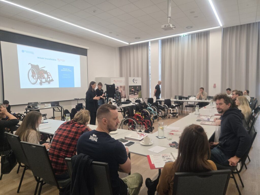 Prezentacja w sali konferencyjnej o wózkach inwalidzkich z wystawionymi modelami i uczestnikami siedzącymi przy stołach.