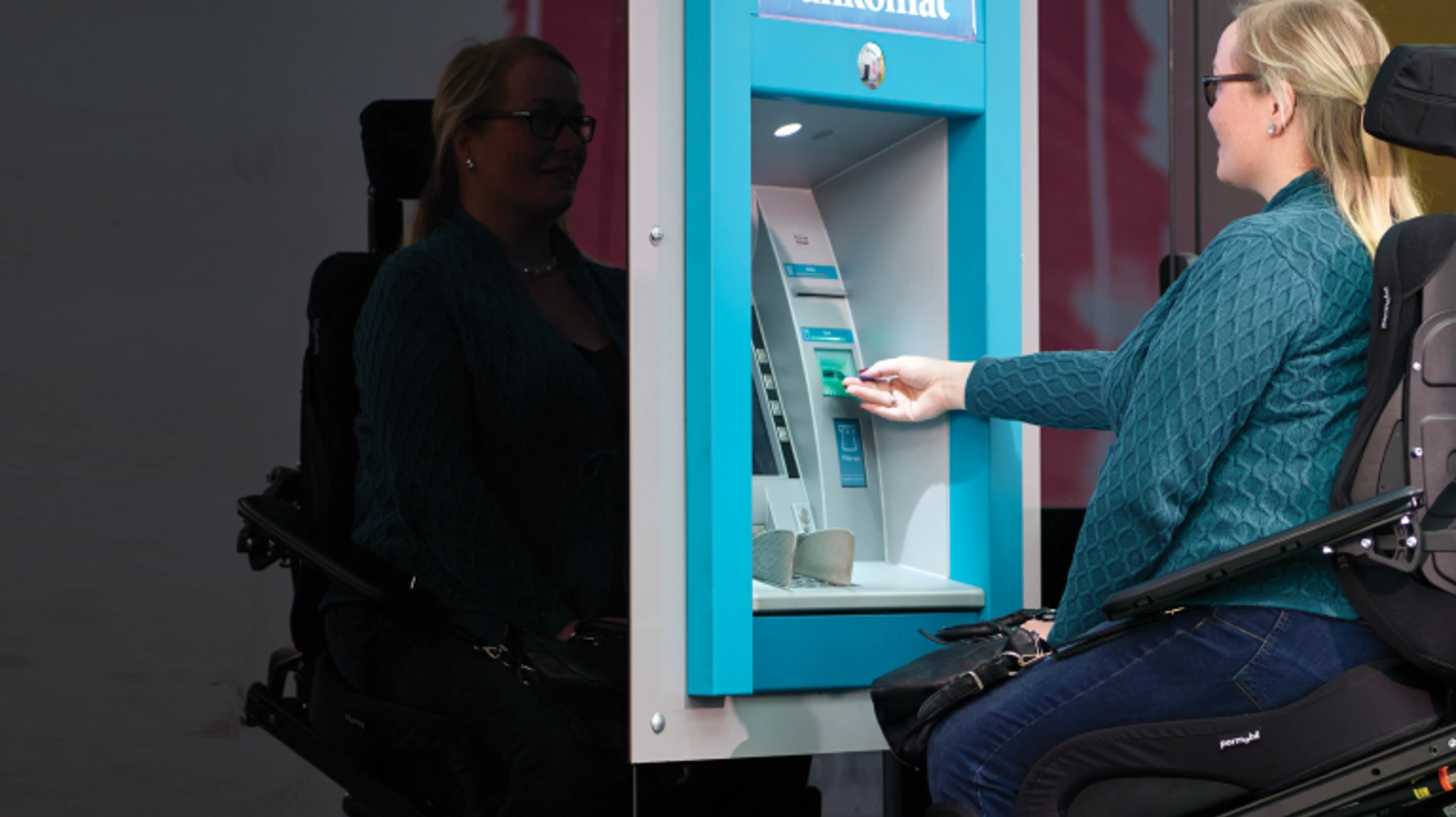 Osoba siedząca na wózku inwalidzkim wkłada kartę do bankomatu o jasnoniebieskiej ramie.
