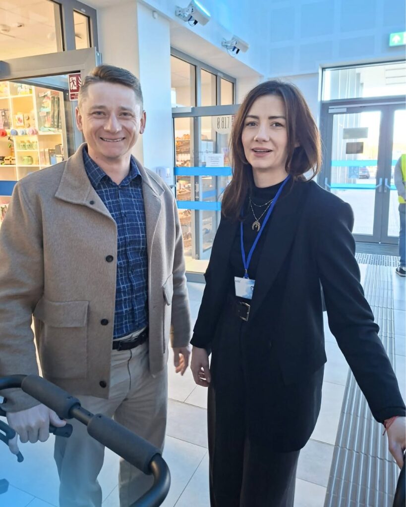 Mężczyzna w beżowym płaszczu i kobieta w czarnym żakiecie z identyfikatorem stoją w jasnym holu przy drzwiach sklepu.
