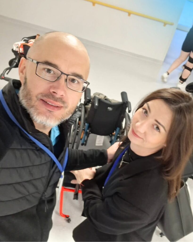 Mężczyzna w okularach i kobieta robią selfie na korytarzu przed kilkoma wózkami inwalidzkimi, oboje z niebieskimi identyfikatorami.