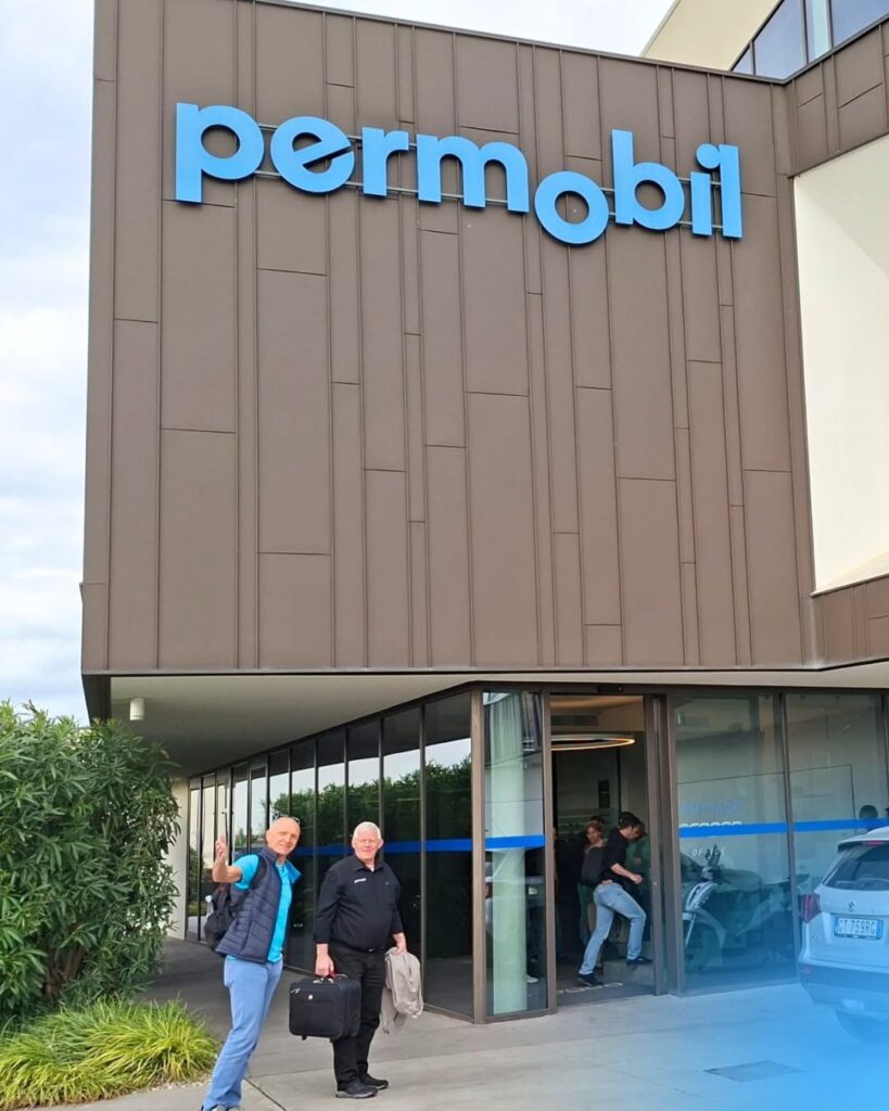 Budynek z dużym niebieskim logo permobil nad przeszklonym wejściem, przed którym stoją dwaj mężczyźni z bagażem.