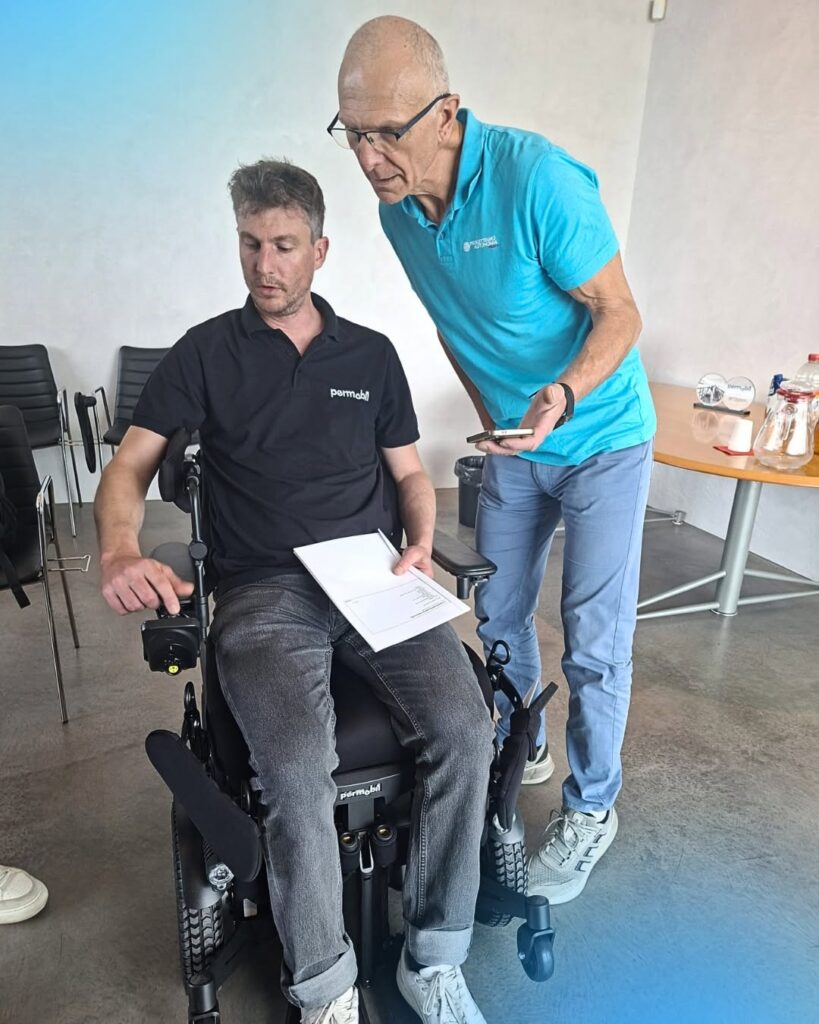 Mężczyzna w elektrycznym wózku inwalidzkim obsługuje joystick, a stojący obok mężczyzna w turkusowej koszulce pokazuje mu telefon.