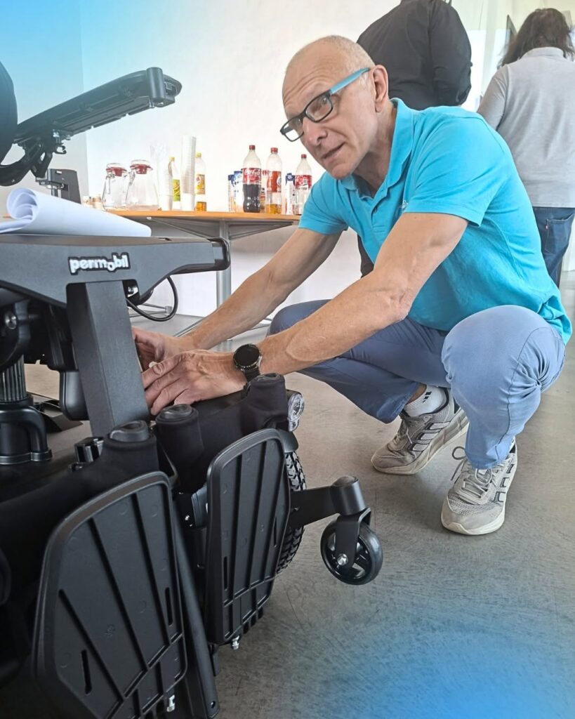 Mężczyzna w turkusowej koszulce i okularach kuca i reguluje mechanizm elektrycznego wózka inwalidzkiego marki Permobil, stół z napojami w tle.