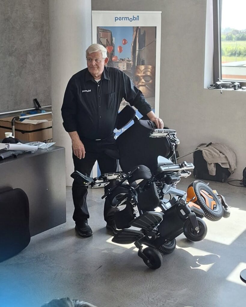 Starszy mężczyzna w czarnej koszuli z logo Permobil stoi obok złożonego elektrycznego wózka inwalidzkiego w biurze.