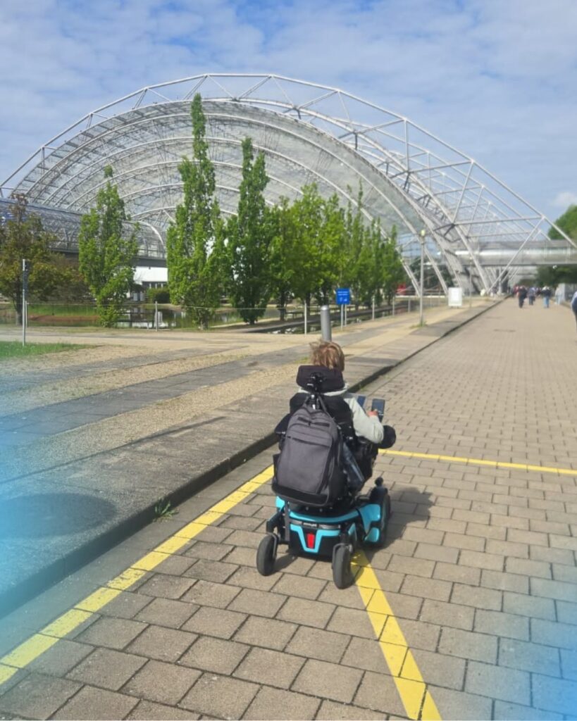 Osoba na elektrycznym wózku inwalidzkim z plecakiem jedzie chodnikiem w stronę dużej szklano-stalowej, łukowej hali.
