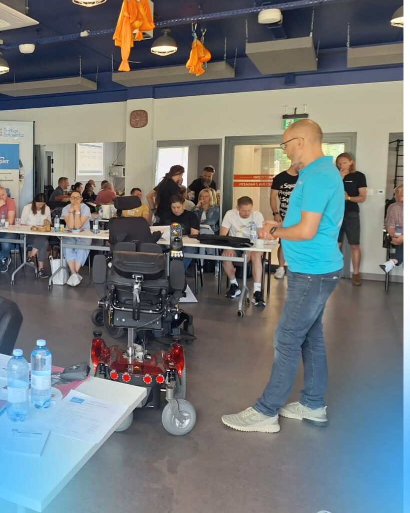 Mężczyzna w turkusowej koszulce prezentuje elektryczny wózek inwalidzki przed grupą osób w sali warsztatowej.